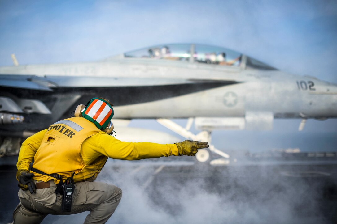 A sailor extends his arm to signal to an aircraft to launch from a ship's deck.