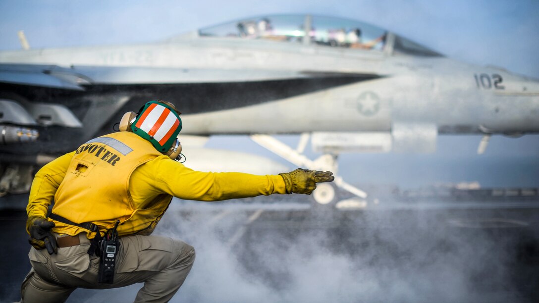 A sailor extends his arm to signal to an aircraft to launch from a ship's deck.