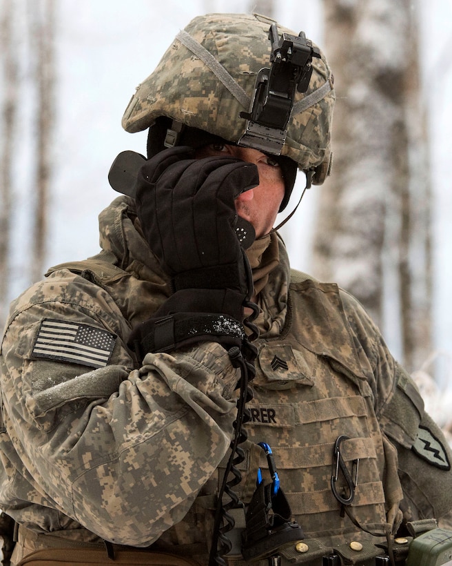 A soldier speaks into a radio he's holding with a black-gloved hand outside in the snow.