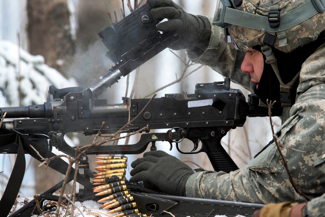 A soldier in a prone position clears an M240B machine gun in snowy, wooded terrain.