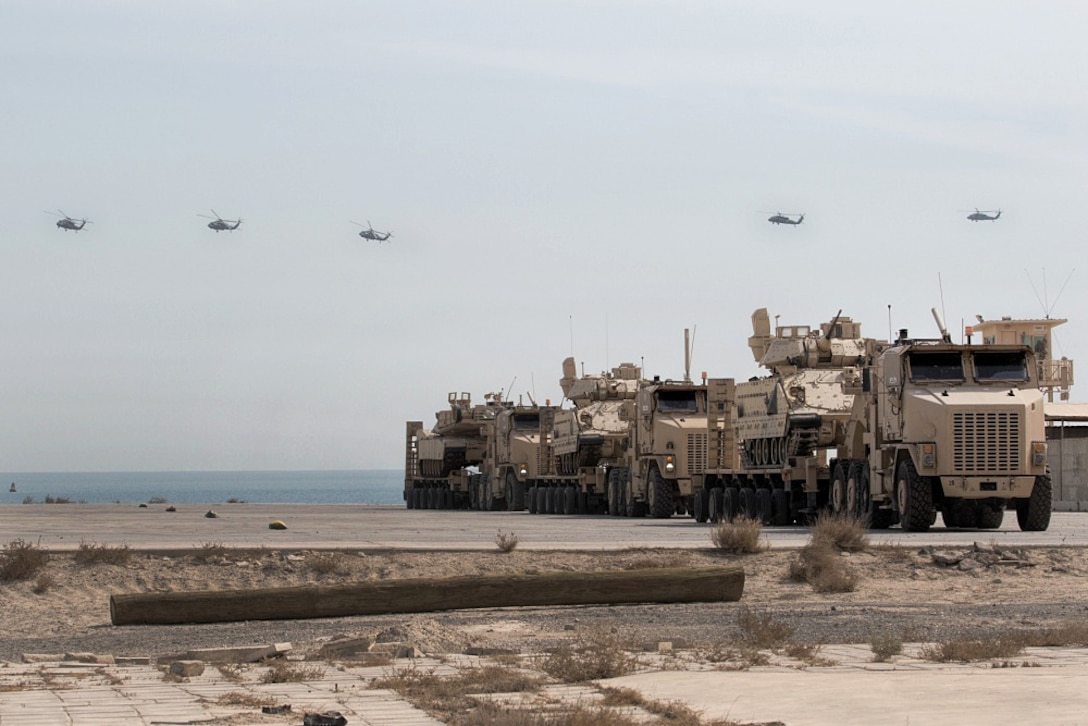 UH-60 Black Hawk helicopters fly over armored vehicles that have been loaded onto trucks.