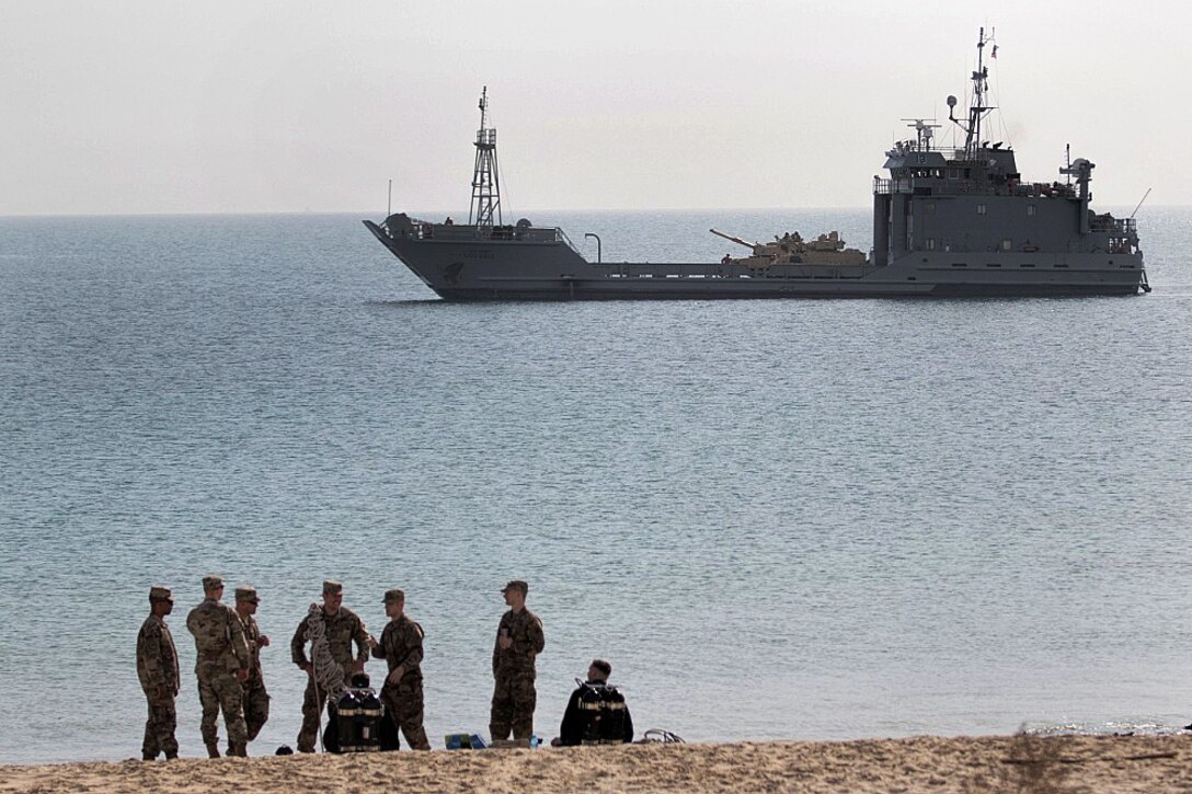 Army divers stand on shore as a ship sits in the water off shore.