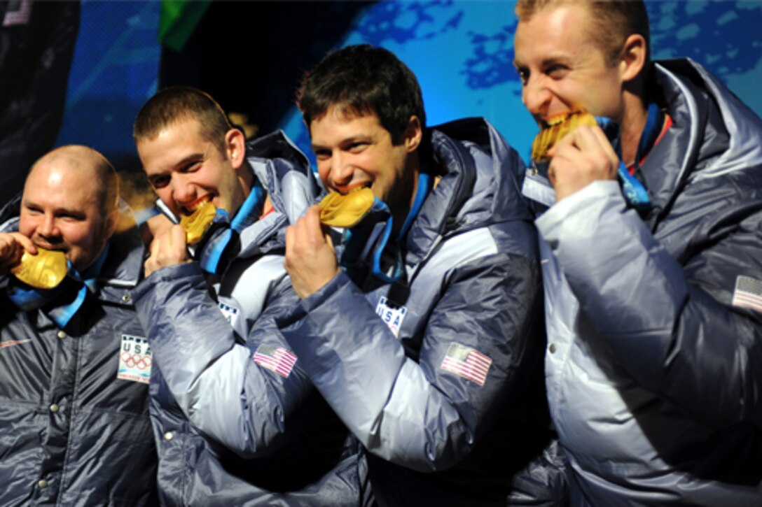 Four men pose with Olympic gold medals in their mouths.
