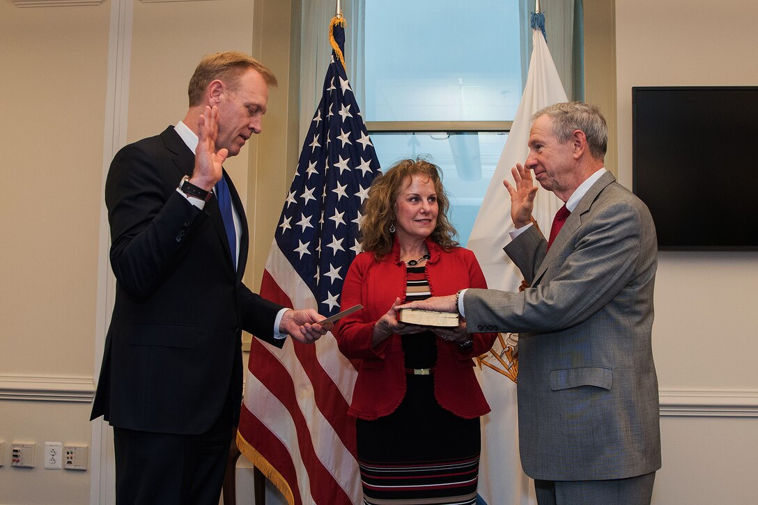 The deputy defense secretary raises his right hand while another person lays his hand on a book and raises his right hand.