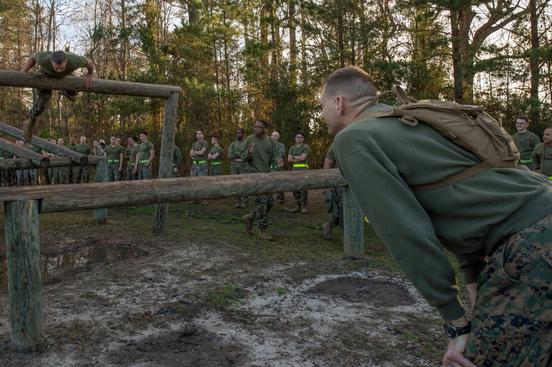 A Marine bends forward to watch another Marine climb over a wooden beam.