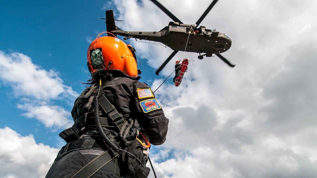 An Army helicopter lifts a red stretcher as a rescue technician guides it.