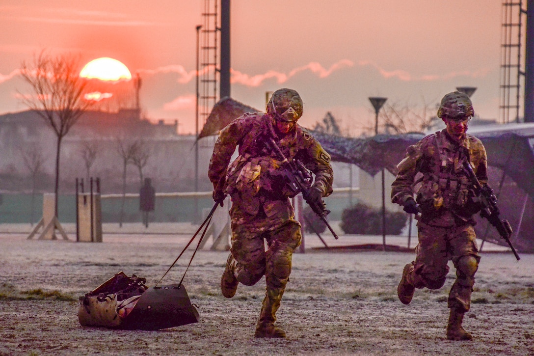 Two soldiers run during an event to earn an expert badge as the sun glows from the horizon.