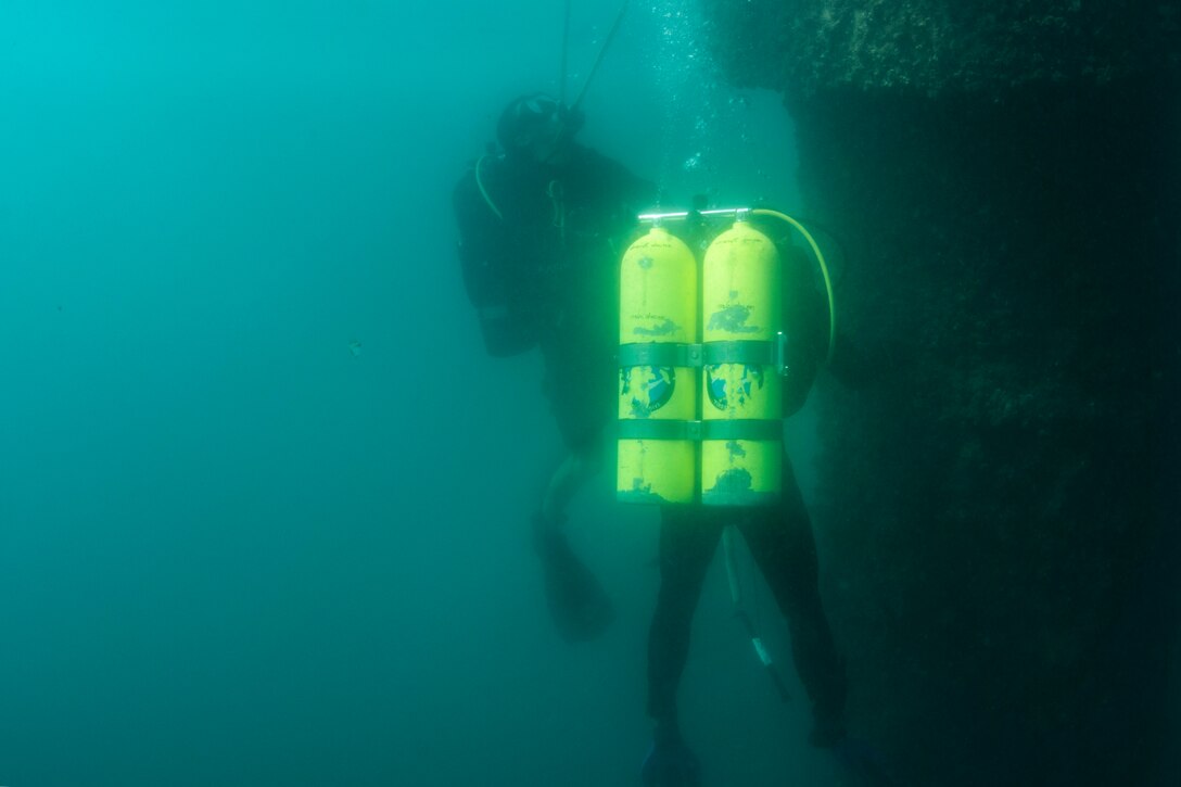 A sailor with yellow oxygen tanks inspects a pier underwater.