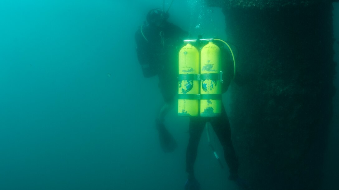 A sailor with yellow oxygen tanks inspects a pier underwater.