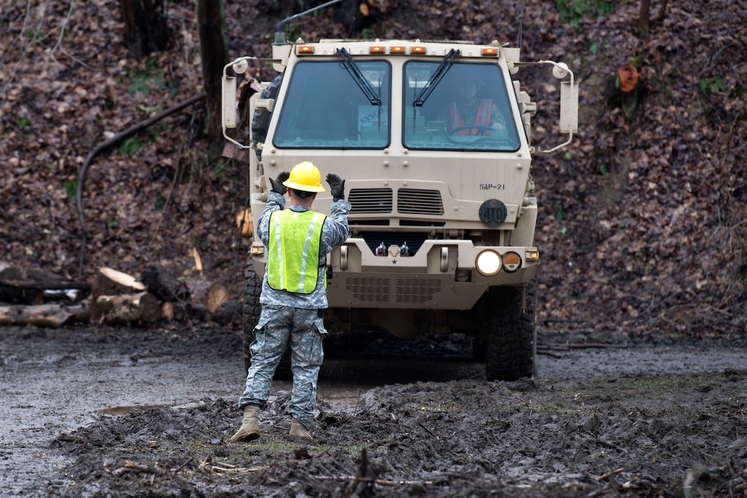 A soldier uses hand signals to direct a truck though water.