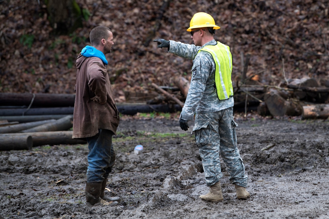 A soldier talks to a local resident while standing in a muddy area.