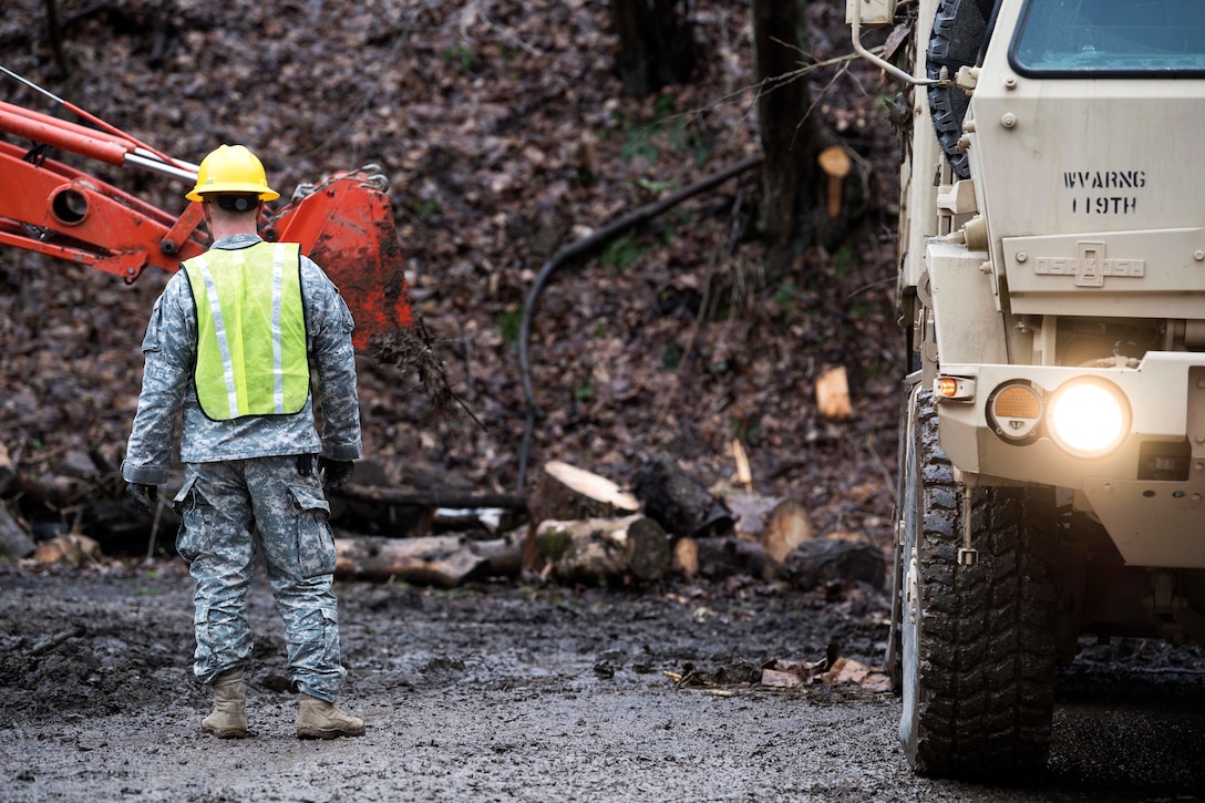 A soldier stands between heavy construction equipment and a truck.