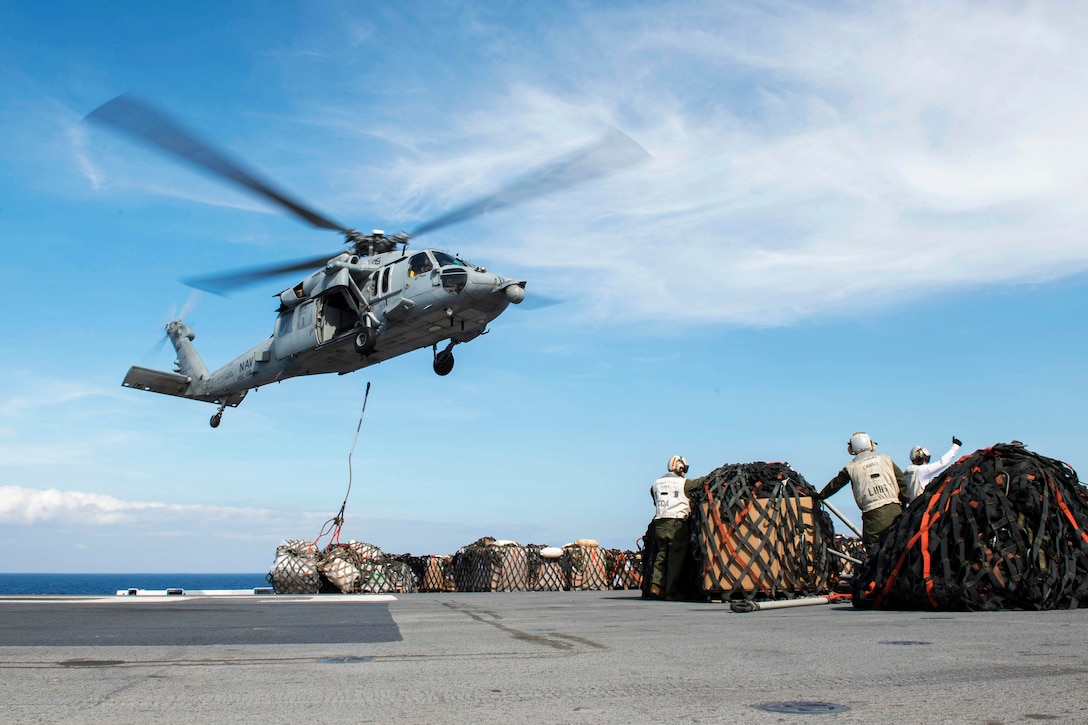 An MH-60S Seahawk hovers over a flight deck with supplies.