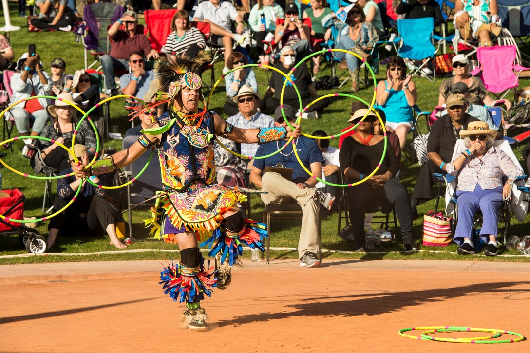 A man in Native American garb holds multiple hoops while dancing in front of a crowd outside on a lawn.