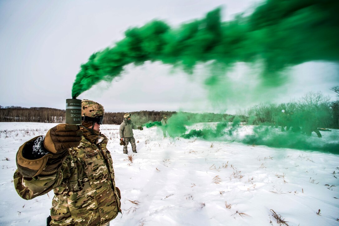 Green smoke streams from a can an airman holds up while standing in snowfield.