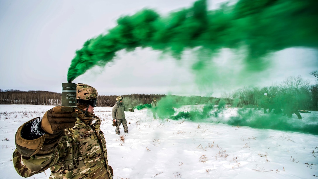 Green smoke streams from a can an airman holds up while standing in snowfield.