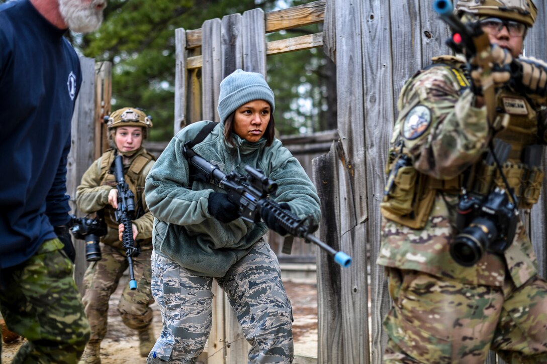 Airmen wielding rifles move forward beside a wood wall.