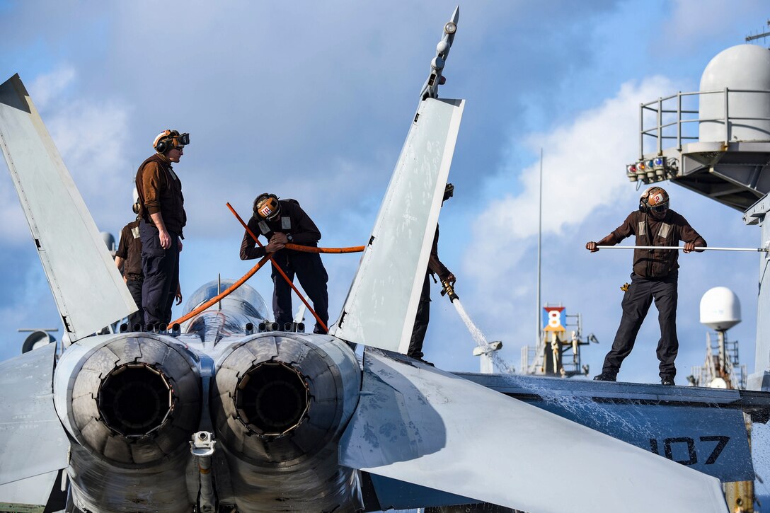 Four sailors stand atop an aircraft and wash it using a hose and mops.