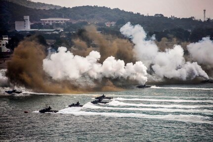 Amphibious vehicles travel in water toward a beach as white and brown smoke wafts in the background.
