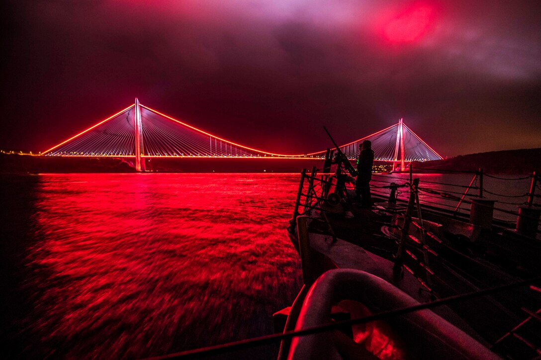 Sailors man a gun on a ship and look out over a suspension bridge illuminated in red light at night illuminated in red light.