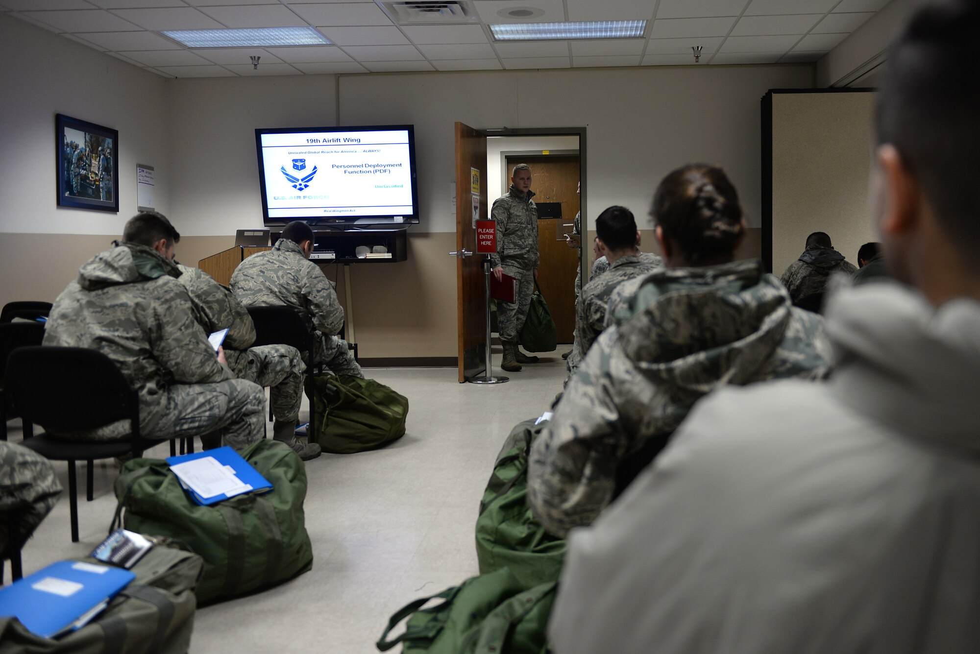 Airmen sit in a room with deployment bags on the ground next to them watching a screen.