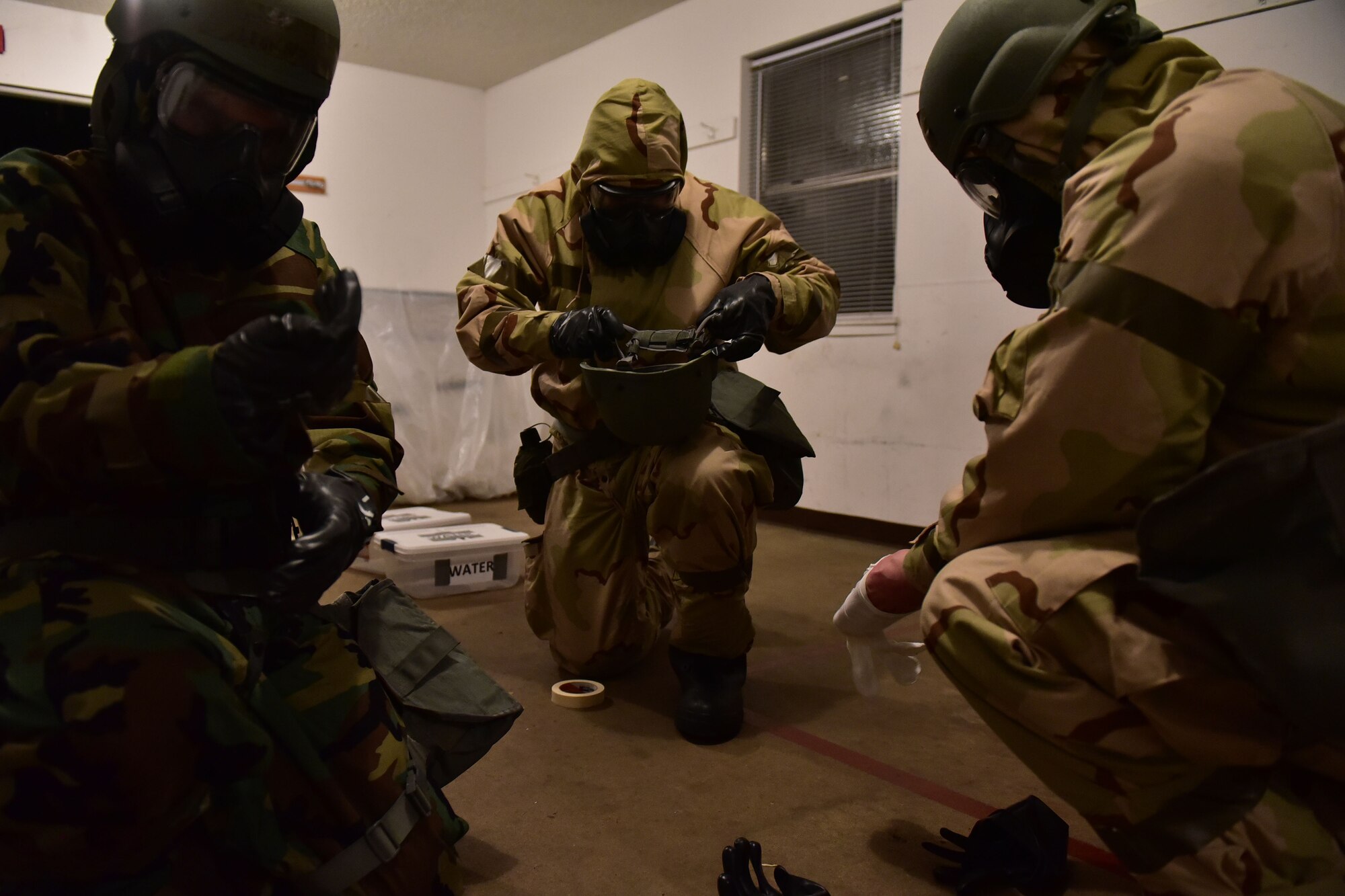 Three Airmen kneel and equip MOPP gear.