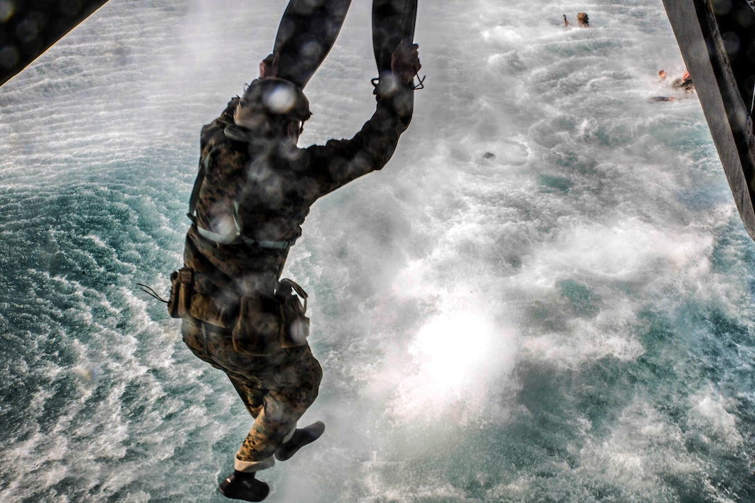A Marine with flippers on his hands raises his arms while leaping from a helicopter over choppy waters.