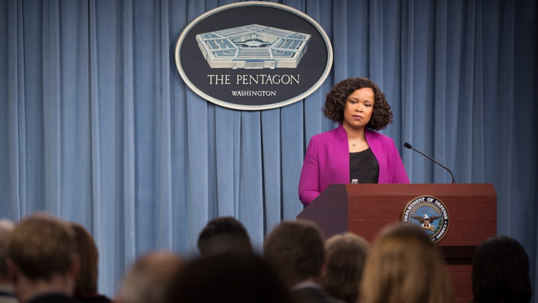 A civilian stands behind a podium in front of a seated audience.