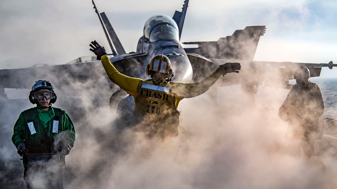 An sailor holds his arms out in front of an aircraft as smoke wafts upward around his waist.