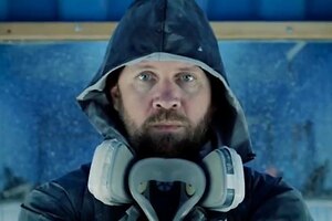 A bearded man wearing a hooded windbreaker stares ahead of him in front of a blue wall.