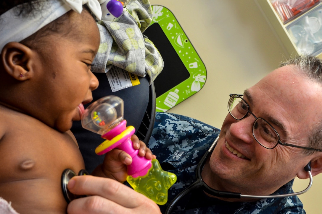 A doctor smiles while listening the a smiling baby's heartbeat with a stethoscope.