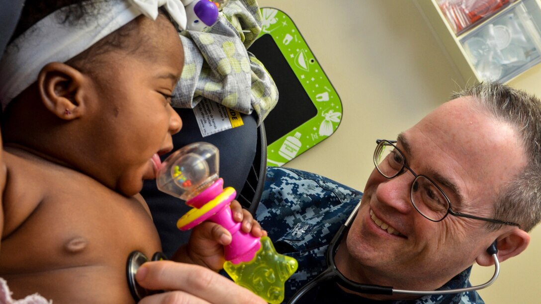 A doctor smiles while listening the a smiling baby's heartbeat with a stethoscope.