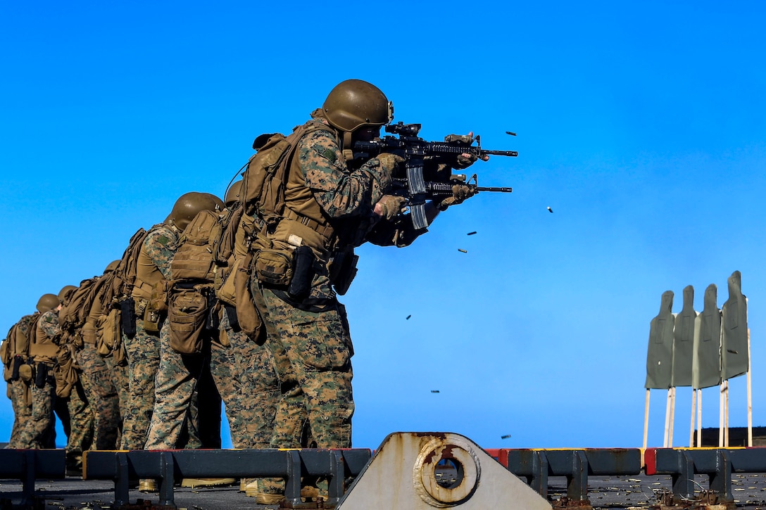 Marines stand in a row and fire rifles at targets on a ship's deck against blue skies.
