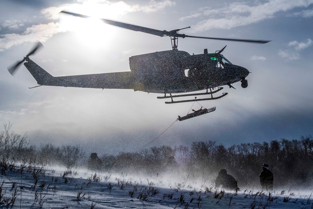 Snow whips up from a field as a helicopter transports a person lying on a stretcher by a sling load.