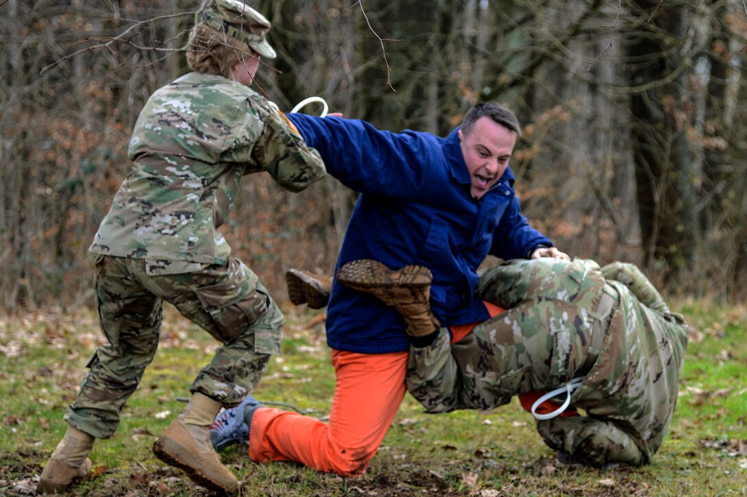 Two soldiers struggle with a soldier in civilian clothing in a field outside.