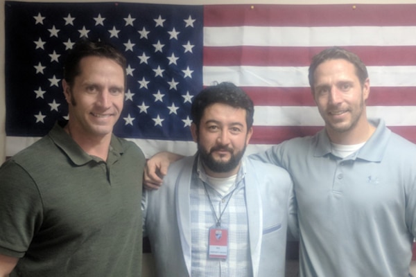 Three men pose for a photograph in front of an American flag in Afghanistan.