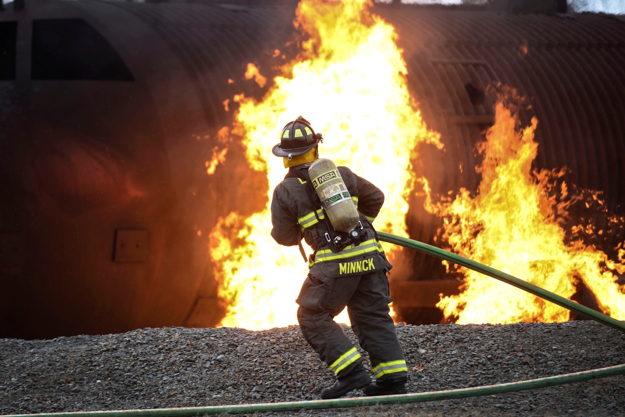 A male firefighter uses a firehose to extinguish a fire during a training burn on a simulated aircraft.