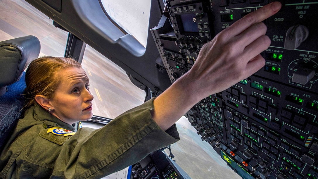 A pilot sitting in a cockpit raises her arm while manipulating something on the aircraft's control panel.