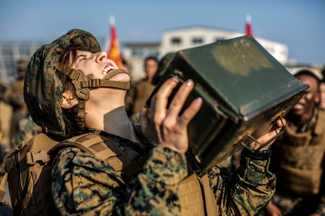 A Marine closes her eyes and tilts her face upward while trying to lift an ammo can, as fellow troops watch.