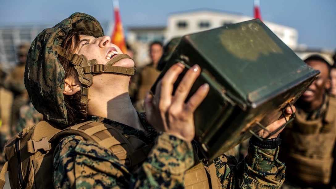 A Marine closes her eyes and tilts her face upward while trying to lift an ammo can, as fellow troops watch.