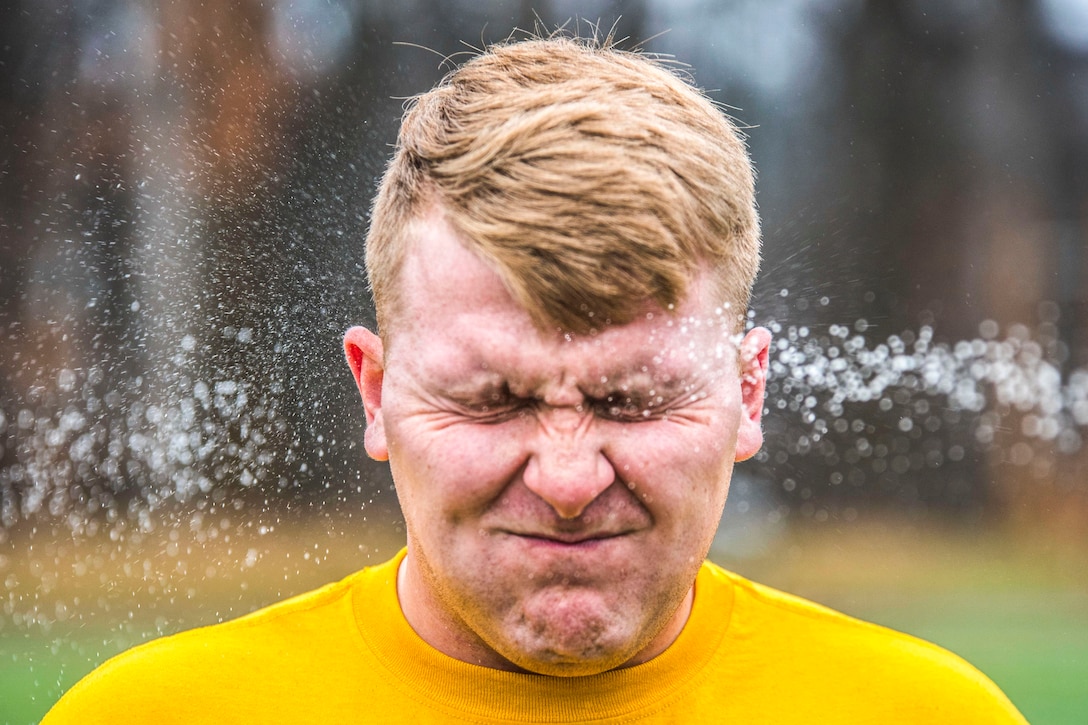 A sailor in a yellow t-shirt holds his eyes and mouth closed as a spray of liquid hits him.