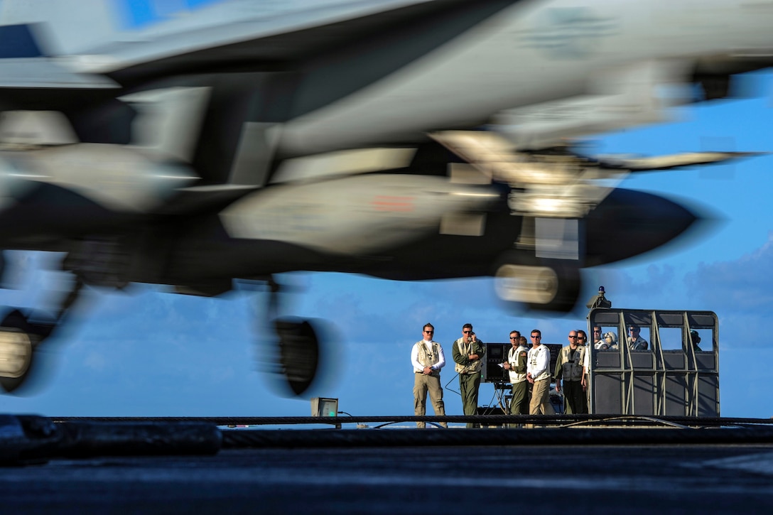 Sailors watch as a jet lands on a flight deck.