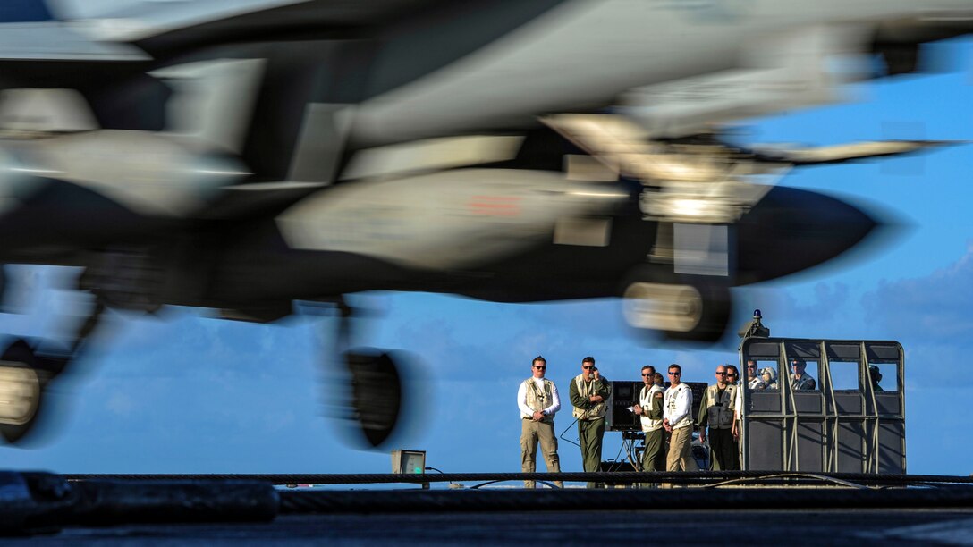 Sailors watch as a jet lands on a flight deck.