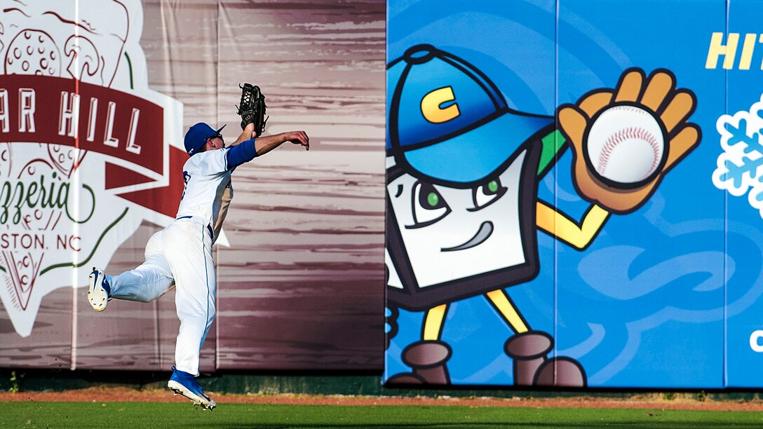 A baseball player catches a ball against a wall with a cartoon illustration of a home plate character catching a ball.