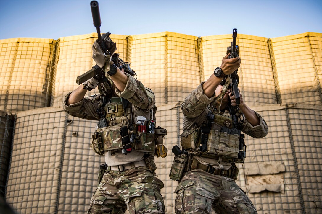 Airmen point rifles, which obscure their faces, toward the camera while standing outside in front of a military barrier.