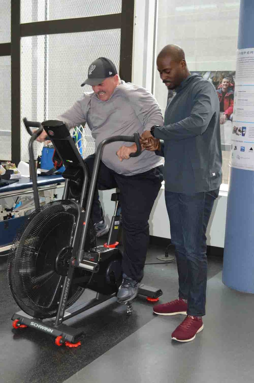 Man rides exercise bike while physical therapist watches.