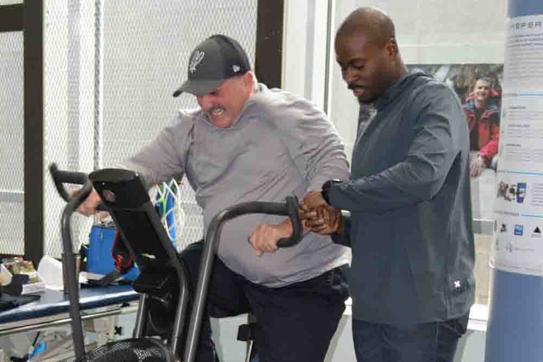 A man rides an exercise bike while a trainer stands next to him.
