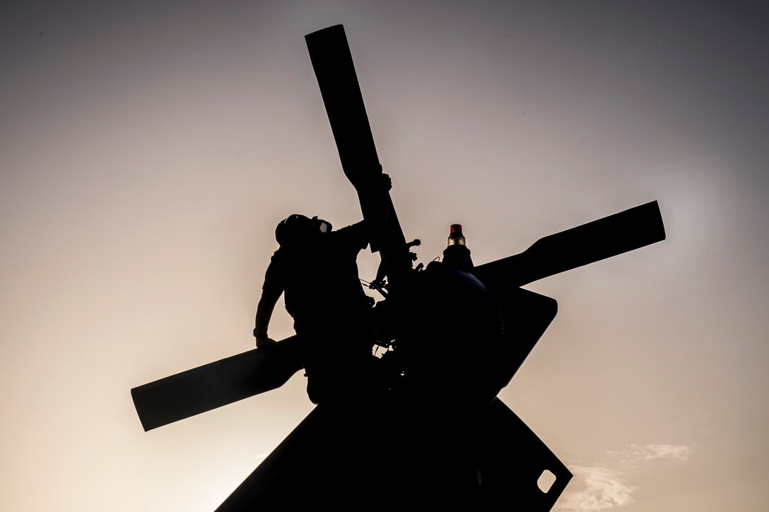 A sailor, shown in silhouette, inspects a helicopter's tail rotor.