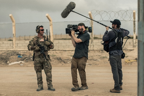 A man holding a camera and a man holding a large mic talk to a service member.