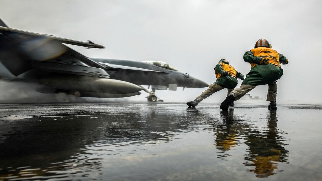 Two sailors, shown from behind, lean over and signal as a jet takes off from a rainy flight deck.